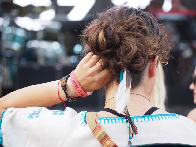 Back view of relaxed woman with feathers in hair attending outdoor music festival. Wristbands on her wrist indicating entry to event. Can be used to promote festivals, summer events, concerts, and boho-style fashion.