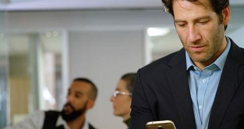 caucasian businessman engrossed in smartphone tasks, standing amidst busy office activity. two colleagues, slightly blurred in background, emphasizing teamwork and a dynamic work setting. suitable for themes of corporate lifestyle, modern technology usage, or illustrating a professional environment.