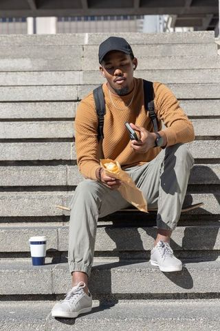 Young african american man sitting on steps holding smartphone eating snack wearing earbuds