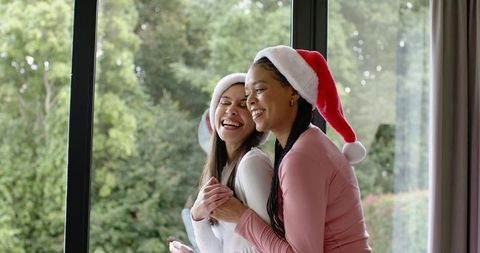 Two friends wearing santa hats hugging and laughing by window overlooking garden