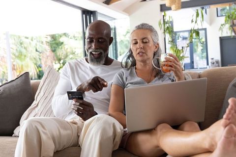 Mature multiracial couple shopping online from living room with laptop and credit card