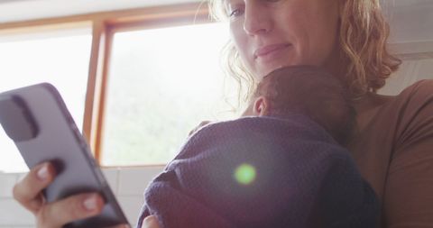 Mother bonding with newborn while checking smartphone in kitchen