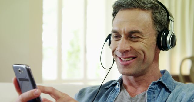 A middle aged man wearing headphones while using his smartphone, enjoying music. Ideal for use in website banners, promotional materials for music streaming services, technology gadgets, and lifestyle blogs focused on digital entertainment.
