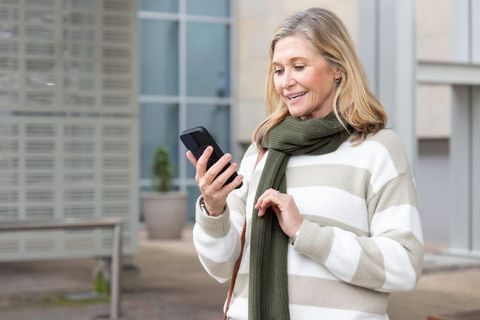 Smiling senior woman checking smartphone outdoors in urban courtyard wearing green scarf