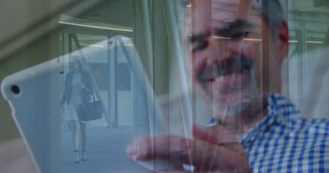 Smiling mature man tapping tablet with female commuter reflection in modern glass lobby