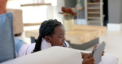 African american girl lying on sofa holding tablet, using device in bright modern room