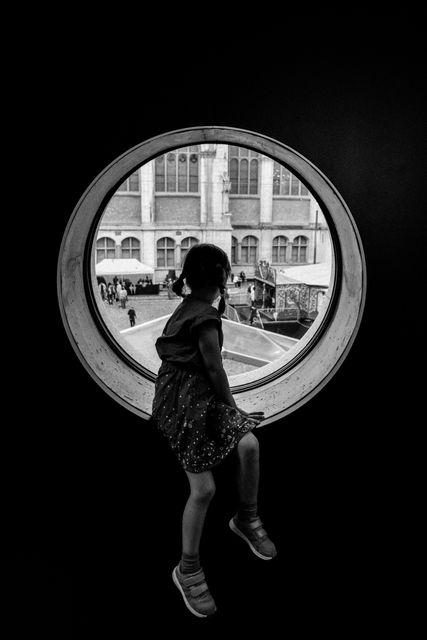 Child sitting on windowsill, viewing a courtyard scene through a circular window in a museum. Black and white photography captures a sense of curiosity and wonder. Useful for themes related to childhood, curiosity, architecture, or indoor activities.