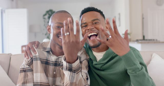 Happy diverse gay male couple embracing on couch and showing wedding rings at home. Togetherness, relationship, love and domestic life, unaltered.