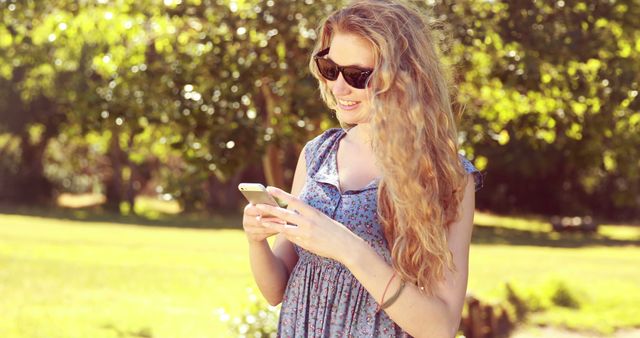 A woman with long hair is smiling while texting on her smartphone in a sunny park. She is wearing a casual dress and sunglasses, suggesting a relaxed and laid-back moment enjoying nature. Perfect for advertising outdoor activities, mobile technology, lifestyle, and fashion.