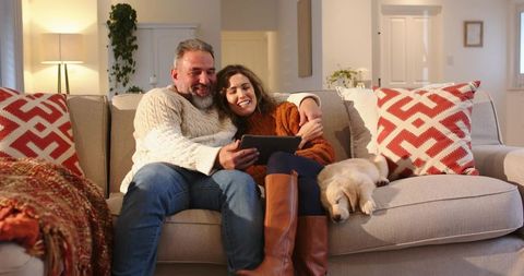 Middle-aged couple cuddling on couch using tablet with sleeping puppy cozy home moment