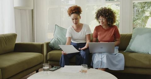 African American women reviewing documents on sofa with laptop in sunlit living room