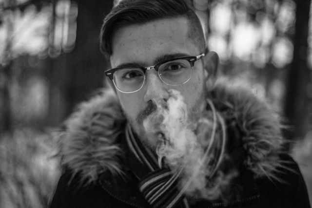 Young man with glasses is exhaling vapor in a winter forest. He is dressed in a fur-collared coat and a striped scarf. The monochrome setting adds a dramatic and moody effect, highlighting the cold weather. Perfect for illustrating winter fashion, outdoor activities, or emotions evoked by winter.