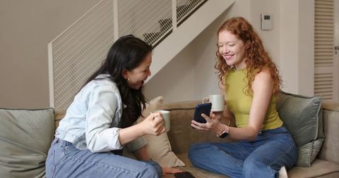 Sharing coffee and laughter on sofa while checking smartphone, casual cozy home friendship