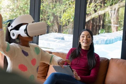 Mother watching child playing with VR headset while holding tablet on living room couch