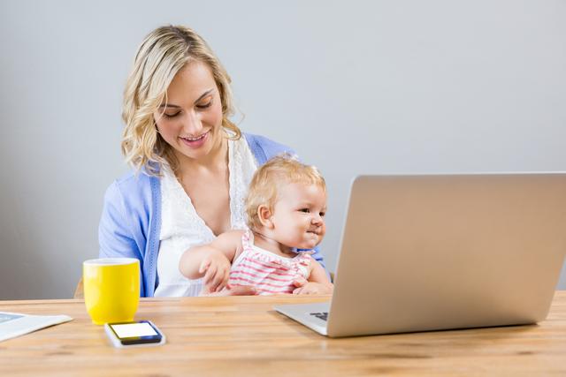 Young mother holding baby girl while using laptop at home. Ideal for articles on work-life balance, remote work, parenting tips, and family life. Can be used in blogs, websites, and advertisements promoting flexible work arrangements, parenting products, or family-friendly workplaces.