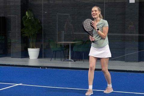 Smiling young woman playing padel on blue court wearing smartwatch and tennis skirt