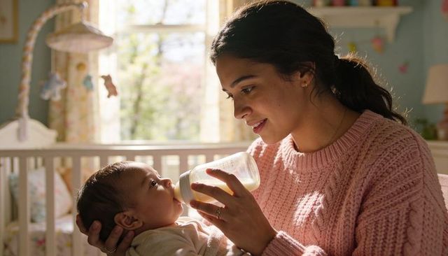 Mother feeding infant with bottle in nursery, cradling in pink sweater, warm morning light
