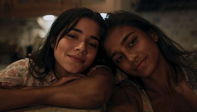 Two women leaning heads on kitchen counter under warm cabinet lights, intimate closeup