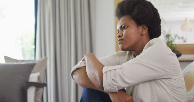 Woman sitting by window in cozy living room during the day, looking thoughtfully outside. Could be used for depicting introspection, mental health awareness, or concepts of relaxation and quiet moments at home. Perfect for lifestyle blogs, therapy websites, or home decor inspiration.