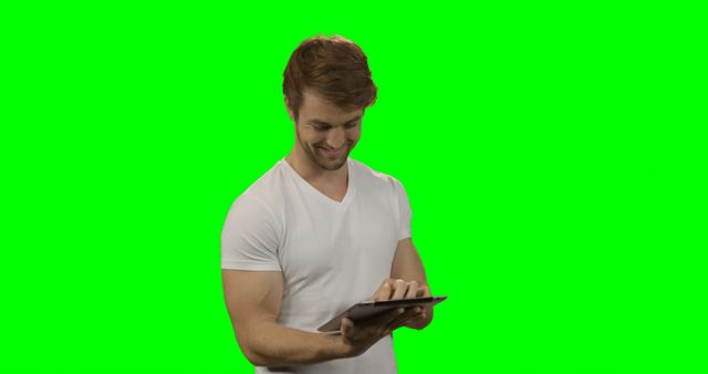 Young man standing and using a tablet with a green screen background. He is dressed in a casual white shirt and is smiling while interacting with the device. This can be used in tech-related ads, digital marketing materials, and web design projects to emphasize modern technology and interactivity.