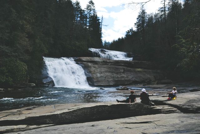 People relaxing near a waterfall in a forested area. Ideal for use in travel blogs, nature magazines, relaxation and wellness promotions, and eco-tourism advertisements. Captures the natural beauty and serenity of outdoor environments, promoting the allure of nature and adventure.