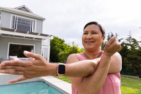 Mature woman stretching at poolside, wearing smartwatch and pink top, smiling outdoors