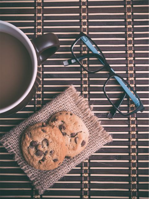 Close-up of coffee cup, black-rimmed glasses, and two chocolate chip cookies placed on a piece of burlap over a bamboo mat surface. Perfect for themes related to cozy mornings, coffee time, breakfast, relaxation, and rustic setups. Ideal for use in blog posts, social media, breakfast recipes, snack ideas, and lifestyle magazine spreads.
