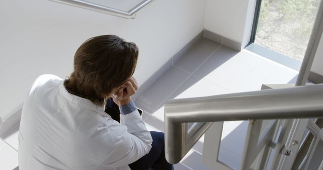 Male doctor in a lab coat sitting alone on a modern staircase, looking pensive and reflective. This serene, thoughtful setting highlights the stresses and solitude that can accompany a medical profession. Ideal for articles or blogs discussing mental health, healthcare, the challenges faced by medical professionals, or work-life balance.