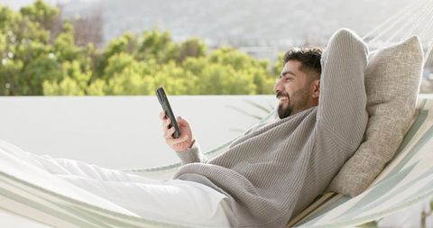 Indian man lounging in hammock on sunny terrace smiling while using smartphone