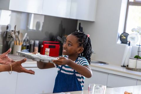 Young girl offering muffin tray while baking with parent in bright modern kitchen
