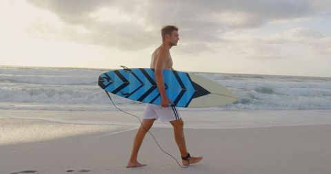 Young Man Strolling on Beach Holding Surfboard During Sunrise