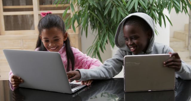 Two children sitting at black table using laptops and smiling indoors. Great for education, technology, teamwork, and diversity concepts.