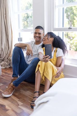 Laughing diverse couple taking selfie by sunlit windows in cozy hardwood-floor home