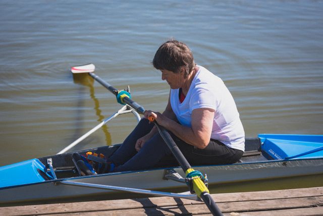 Senior woman sitting in a rowing boat on the river, preparing for a rowing session. Ideal for use in articles or advertisements promoting active lifestyles, senior fitness, retirement hobbies, and water sports. Can also be used in content related to rowing clubs and outdoor activities for seniors.
