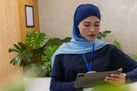 Receptionist wearing two-tone blue hijab holding tablet folio in biophilic lobby