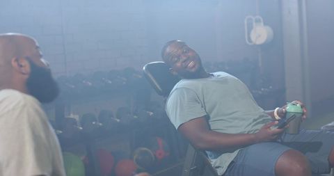 Black male gym partners reclining on incline bench, chatting and holding shaker bottle