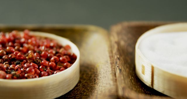 Red peppercorns are displayed in a small wooden bowl next to a bowl of salt on a rustic surface, with copy space. Culinary enthusiasts often use such spices to enhance the flavors in their dishes.
