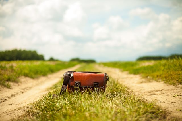 Vintage leather bag placed on a deserted dirt road, surrounded by a rural landscape under a cloudy sky. Ideal for depicting themes of travel, adventure, exploration, solitude, or a rustic lifestyle. Great for travel blogs, adventure stories, and lifestyle magazines.