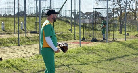 Sunlit baseball teammates practicing in park batting cage holding glove with copy space