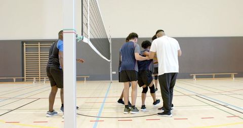 Diverse volleyball teammates supporting injured player in indoor court huddle, coach aiding recovery