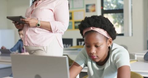 African American Girl Working on Laptop in Sunlit Classroom While Teacher Uses Tablet