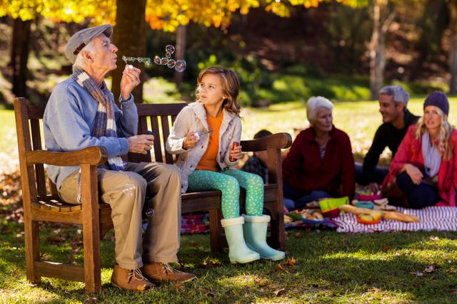 Grandfather and granddaughter enjoying a playful moment blowing bubbles on a park bench during autumn. Family members in the background having a picnic on a blanket. Ideal for illustrating family bonding, outdoor activities, and multigenerational relationships in a natural, autumn setting.