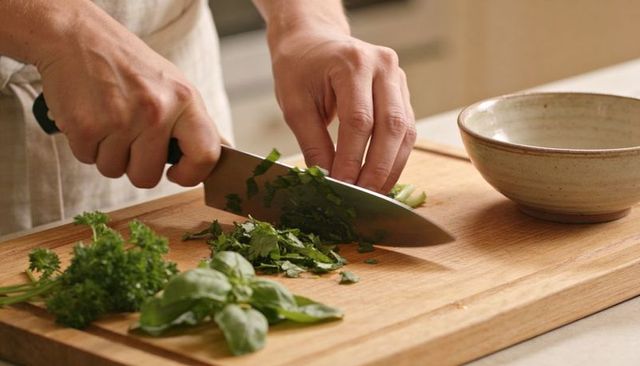 Home chef chopping fresh parsley and basil on wooden cutting board with chef knife