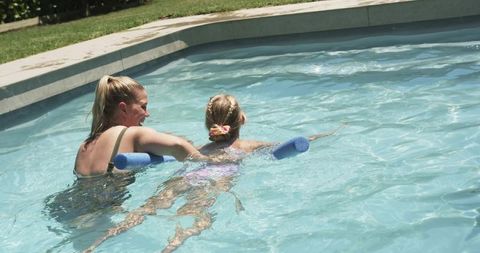 Backyard swimming lesson with instructor guiding child on blue noodle in sunny pool