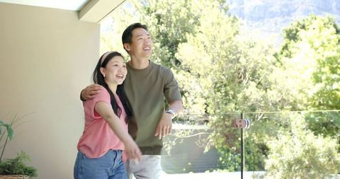 Asian couple enjoying sunny terrace view, smiling and embracing on glass-railed balcony