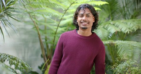 Smiling young asian man standing in greenhouse wearing maroon sweater among ferns