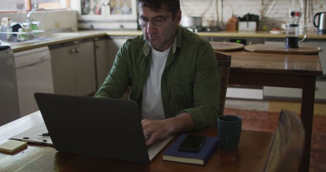 A middle-aged man in casual clothing is working remotely on his laptop while sitting in a home kitchen. There is a coffee mug and a notepad on the table. Ideal for use in articles or blogs about remote work, work-life balance, modern work environments, or productivity tips.