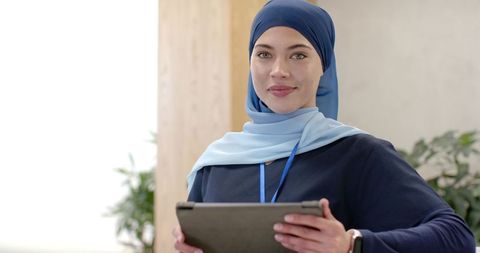 Confident woman wearing blue hijab holding tablet in modern office lobby
