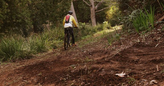A cyclist, clad in sportswear and a helmet, pauses on a dirt trail surrounded by lush greenery, with copy space. Outdoor activities like mountain biking are popular for fitness and connecting with nature.