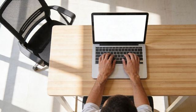 Overhead view man typing laptop at minimalist wooden desk blank screen sunlight remote work